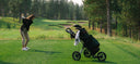Woman swinging a golf club with a golf bag on a golf push cart to the right