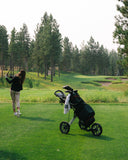 Woman swinging a golf club with a golf bag on a golf push cart to the right
