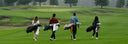 Four people walking on a golf course carrying golf bags, shown from behind in a wide outdoor shot with grass, trees, and sand bunkers in the background.