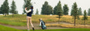 A golfer in a navy vest and beige pants swings a club on a golf course, with a golf bag standing nearby and trees and grass in the background.