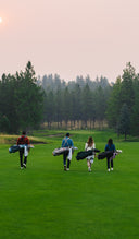 Four people carrying golf bags walk across a green golf course at dusk, with trees and a soft pinkish sky in the background.