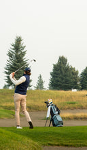 A golfer in a navy vest and beige pants swings a club on a golf course, with a golf bag standing nearby and trees and grass in the background.