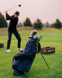 A navy blue golf bag filled with clubs stands on a grassy golf course, with a golfer in a black outfit swinging in the background.