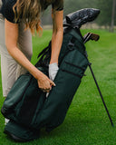 A dark green golf bag on a grassy golf course, with a person bending down to place a golf glove inside the bag.