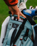 A close-up of a light blue and black golf bag being held by a person on a golf course, with a blue golf club visible behind it.
