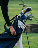 A navy blue golf bag filled with golf clubs and accessories, including white gloves and a headcover with colorful stripes, being carried by a person on a grassy golf course.
