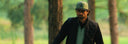 A man wearing a green baseball cap a black quilted vest, and a light green polo shirt walking through a blurred forest of trees.