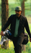 A man wearing a green baseball cap a black quilted vest, and a light green polo shirt walking through a blurred forest of trees.