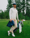 A man on a golf course wearing a white long-sleeve, beige pants, white golf shoes, and a navy cap, holding a blue jacket and a golf bag with clubs.
