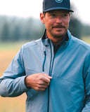 A man wearing a blue zip-up performance jacket and a dark navy baseball cap, standing outdoors in a blurred field setting.