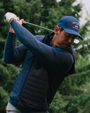 A male golfer in a navy blue zip-up jacket and matching cap swings his club on a golf course