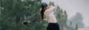 A female golfer in a white long-sleeve top and a blue cap with a white logo is captured mid-swing on a golf course with blurred green trees in the background.
