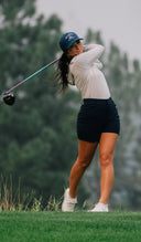 A female golfer in a white long-sleeve top and a blue cap with a white logo is captured mid-swing on a golf course with blurred green trees in the background.