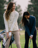 Two women on a golf course, one in a cream zip-up jacket and beige pants preparing to swing, and another in a navy jacket and black pants.