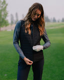 A woman on a golf course wearing a black quilted zip-front vest over a dark long-sleeve shirt and black pants, with a white glove on one hand.