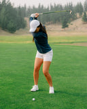 A woman in a navy long-sleeve top, white shorts, and a white cap is captured mid-swing on a golf course