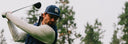 A male golfer in a navy blue cap with a white 'SM' logo, a navy vest, and a light gray long-sleeve shirt is captured mid-swing