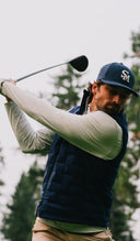 A man in a navy blue quilted vest and matching baseball cap with a white 'SM' logo, captured mid-swing on a golf course.