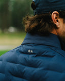 Close-up back view of a person wearing a navy blue puffer jacket and matching cap outdoors, with the small white geometric logo visible on the upper back.