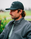 A man in profile wearing a baseball cap with white embroidered text and a gray quarter-zip Kelson jacket
