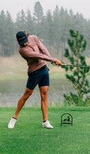 A man wearing a mauve long-sleeve golf shirt, navy blue shorts, and a navy cap is captured mid-swing on a golf course. 