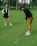 A man in a black long-sleeve shirt, mustard yellow shorts, and white golf shoes putts on a green golf course, while a woman in a white top, navy pleated skirt, and white cap watches from a distance.