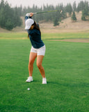 A woman in a navy long-sleeve top, white shorts, and a white cap is captured mid-swing on a golf course
