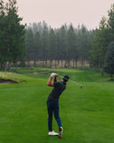 A man in a dark polo shirt, navy pants, and white golf shoes is captured mid-swing on a lush green golf course with a forest in the background.