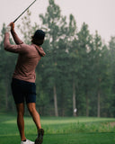 A man wearing a mauve long-sleeve athletic shirt, navy shorts, and a black cap is captured mid-swing on a golf course with trees in the background.