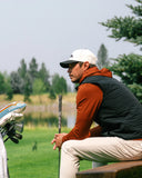 A man on a golf course wearing a white cap, a rust-orange hoodie and black vest, beige pants, sitting on a bench with a golf club and golf bags nearby.