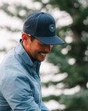 A man wearing a navy blue baseball cap with a circular 'SM' logo on the front and a light blue collared jacket
