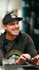 Joel Dahmen wearing a Sun Mountain black baseball cap with white 'SUN.' lettering and a dark long-sleeve shirt with a colorful graphic, seated at a table with cards in a real indoor environment.