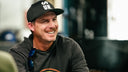 Joel Dahmen wearing a Sun Mountain black baseball cap with white 'SUN.' lettering and a dark long-sleeve shirt with a colorful graphic, seated at a table with cards in a real indoor environment.