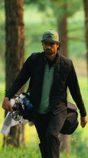 A man walking through a wooded outdoor setting carrying golf clubs and a bag. He is wearing a green cap, a black quilted vest over a light green polo shirt, and dark pants.
