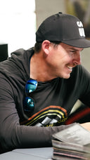 Joel Dahmen wearing a black baseball cap seated indoors with a stack of papers in the foreground.