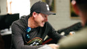 Joel Dahmen wearing a black baseball cap smiling in a casual indoor setting looking at custom bag designs.
