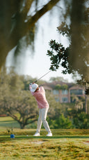 A golfer in a pink shirt and white pants swings a club on a golf course, framed by blurred trees in the foreground.