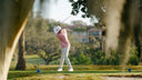 A golfer in a pink shirt and white pants swings a golf club on a sunny golf course, framed by blurred trees in the foreground.
