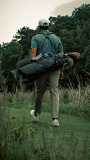 A man seen from behind walking on a grassy golf course while carrying a Matchplay Ballistic golf bag filled with clubs on his shoulder.