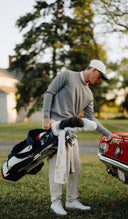Man in a gray sweater and white cap holding a golf bag next to a red car on a grassy area with trees in the background.