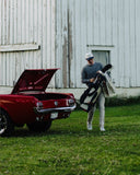 Man with a Hometown golf bag next to an open red car in front of a white wooden building.
