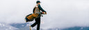 A golfer in a black cap, dark jacket, and beige vest carries a brown and navy golf bag over their shoulder while walking through misty mountains.