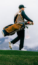 A golfer walking across a grassy hill carrying a brown and navy golf bag with clubs, set against a cloudy sky and distant mountains.
