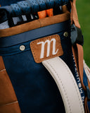 Close-up of a golf bag with navy blue canvas, brown suede panels, and a brown leather patch embroidered with a white script letter 'm'.