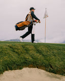 A golfer walks across a grassy golf course slope carrying a large tan and navy golf bag, with a flag and distant hills under an overcast sky.