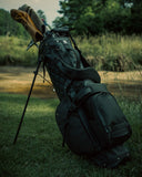 A black Matchplay Ballistic golf bag filled with golf clubs, standing on grass in an outdoor field with trees in the background.