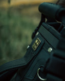 Close-up of a black Matchplay Ballistic logo, shown outdoors against a blurred green and brown background.
