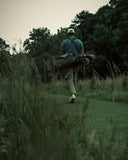 A man walking with a dark green Matchplay Ballistic golf bag with trees and an overcast sky in the background.