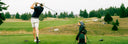 A golfer in a black polo shirt and white shorts swings a club on a grassy golf course, with a green TQR bag on ground next to him