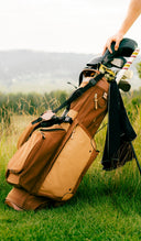 A tan and brown TQR Waxed canvas golf bag filled with clubs, standing on a grassy field with a person's hand reaching for a club